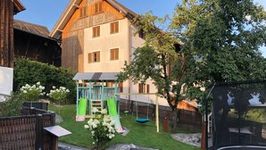 Un jardin avec une terrasse en bois, un trampoline, une aire de jeux et un parterre de fleurs blanches.