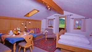 Ein gemütliches Zimmer mit Holzbalkendecke, einem gedeckten Frühstückstisch, einer Couch, einem Bett und Blick auf eine grüne Landschaft durch die Fenster.