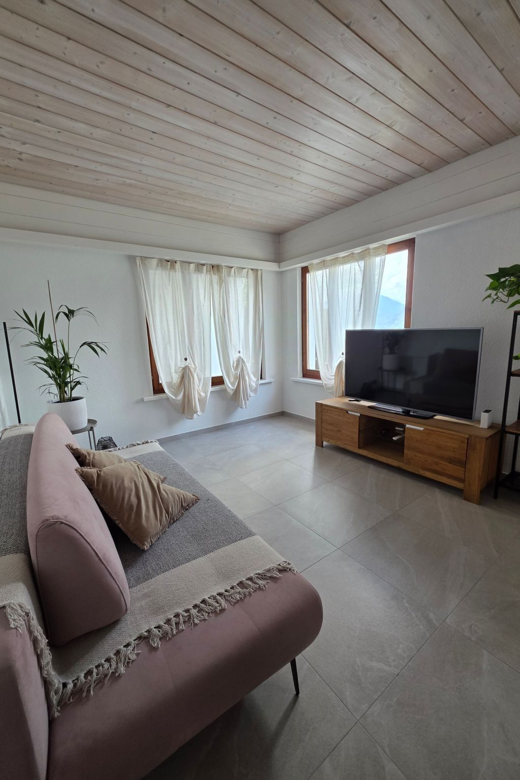 A spacious living room with a large flat-screen TV, wooden ceiling, and large windows with sheer curtains.