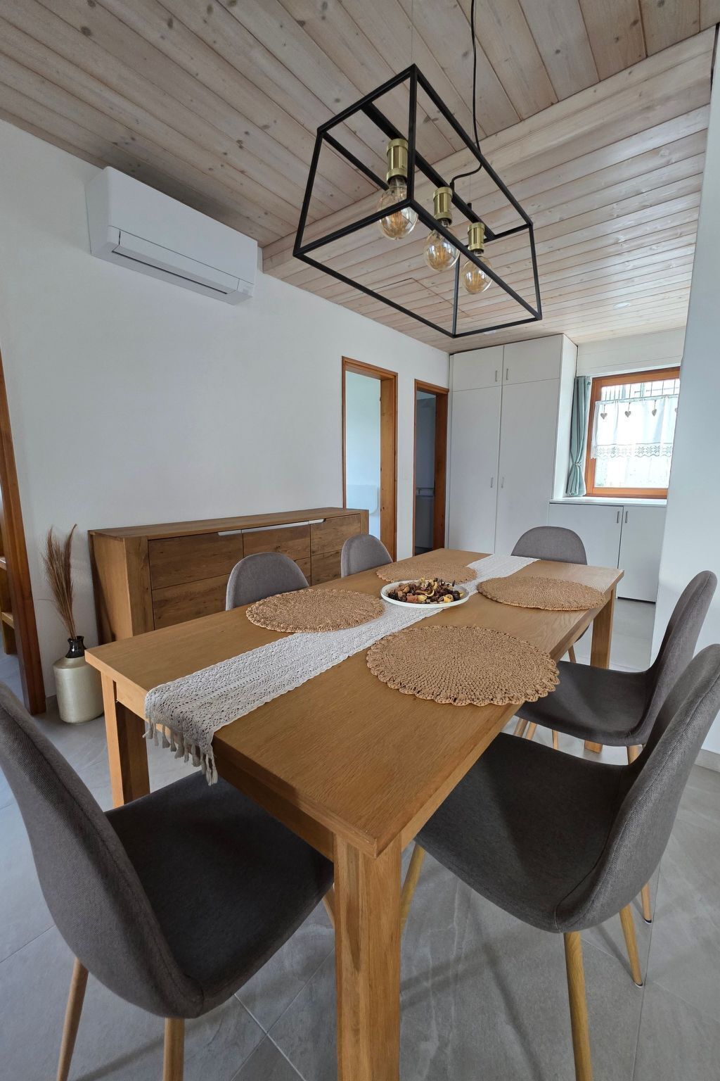 A dining area with a wooden table, four chairs, a decorative ceiling light, and a window with curtains.