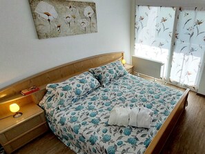 Un dormitorio con cabecera de madera, colcha estampada con motivos florales, mesita de noche con lámpara y ventana con cortinas florales.