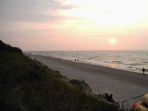 Ein Strand mit Menschen, die am Ufer entlanggehen, einem klaren Himmel und einer untergehenden Sonne im Hintergrund.