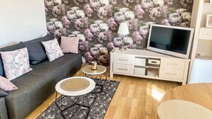 Een woonkamer met een grijze bank, bloemenbehang, een televisie, een lamp, een klein rond tafeltje en een houten vloer.