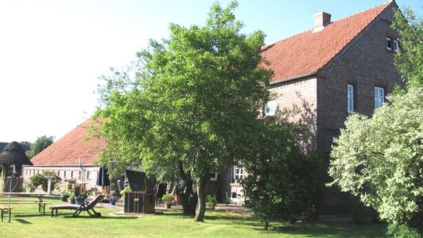 Ein traditionelles Backsteinhaus mit rotem Ziegeldach, umgeben von einem gepflegten Rasen und Bäumen.