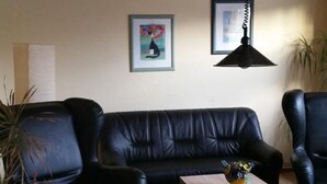 Una sala de estar con un sofá de cuero negro, una mesa de centro de madera y un reposapiés negro. Hay dos cuadros enmarcados en la pared, una planta en maceta y una lámpara colgante.