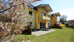 A two-story house with a balcony, a patio with outdoor furniture, and a garden with flowering trees. - Wohnung in Koserow mit Garten und Grill und Neben dem Strand by Interhome (Koserow)