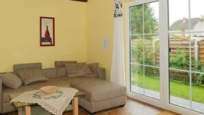A living room with a sectional sofa, a small table with a bowl and a lace tablecloth, and a large sliding glass door leading to a garden. - Vacation home  by Interhome (Bansin)