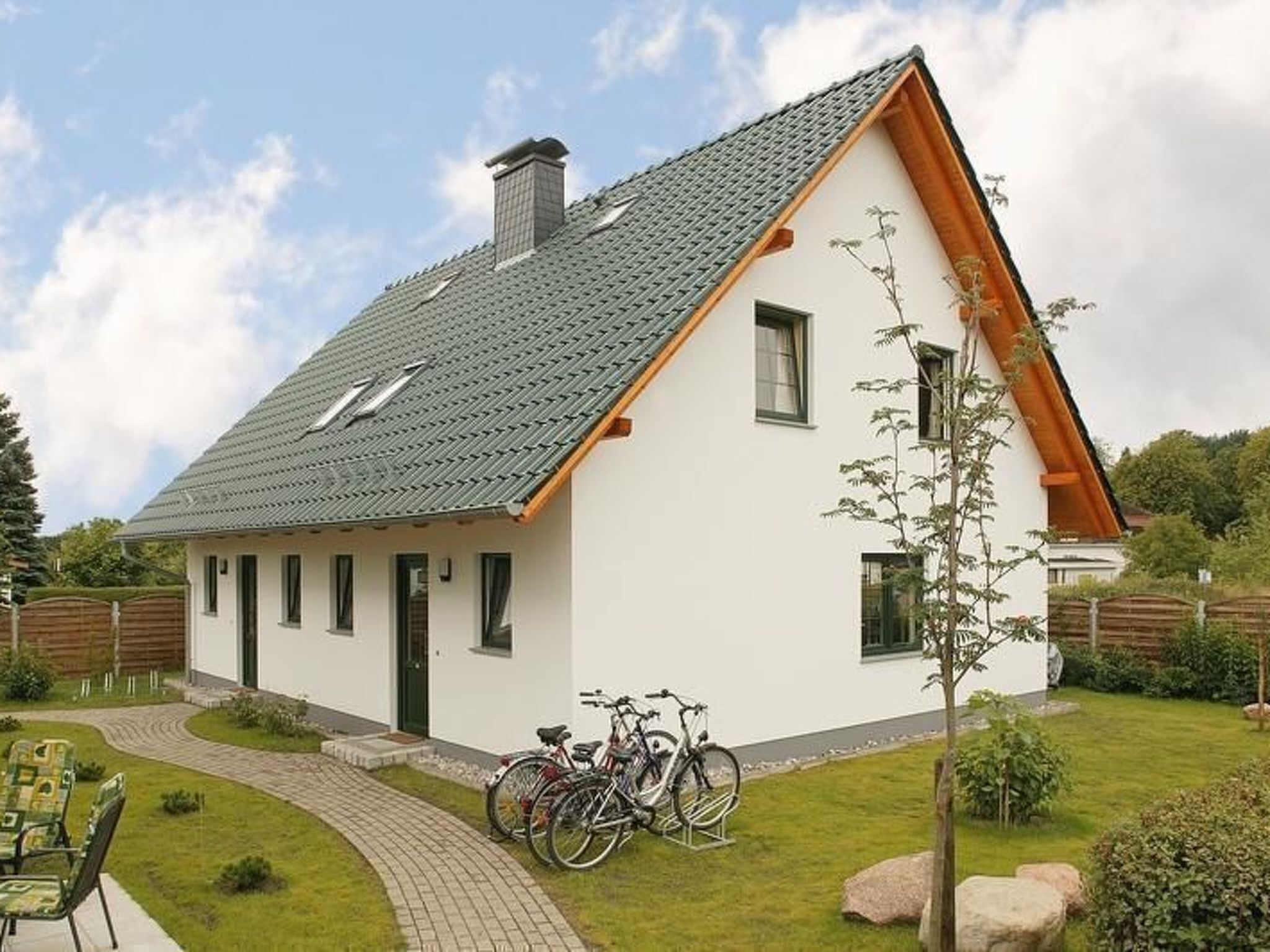 A single-story house with a sloped roof, a visible chimney, and a small garden with a paved pathway and two bicycles parked outside.