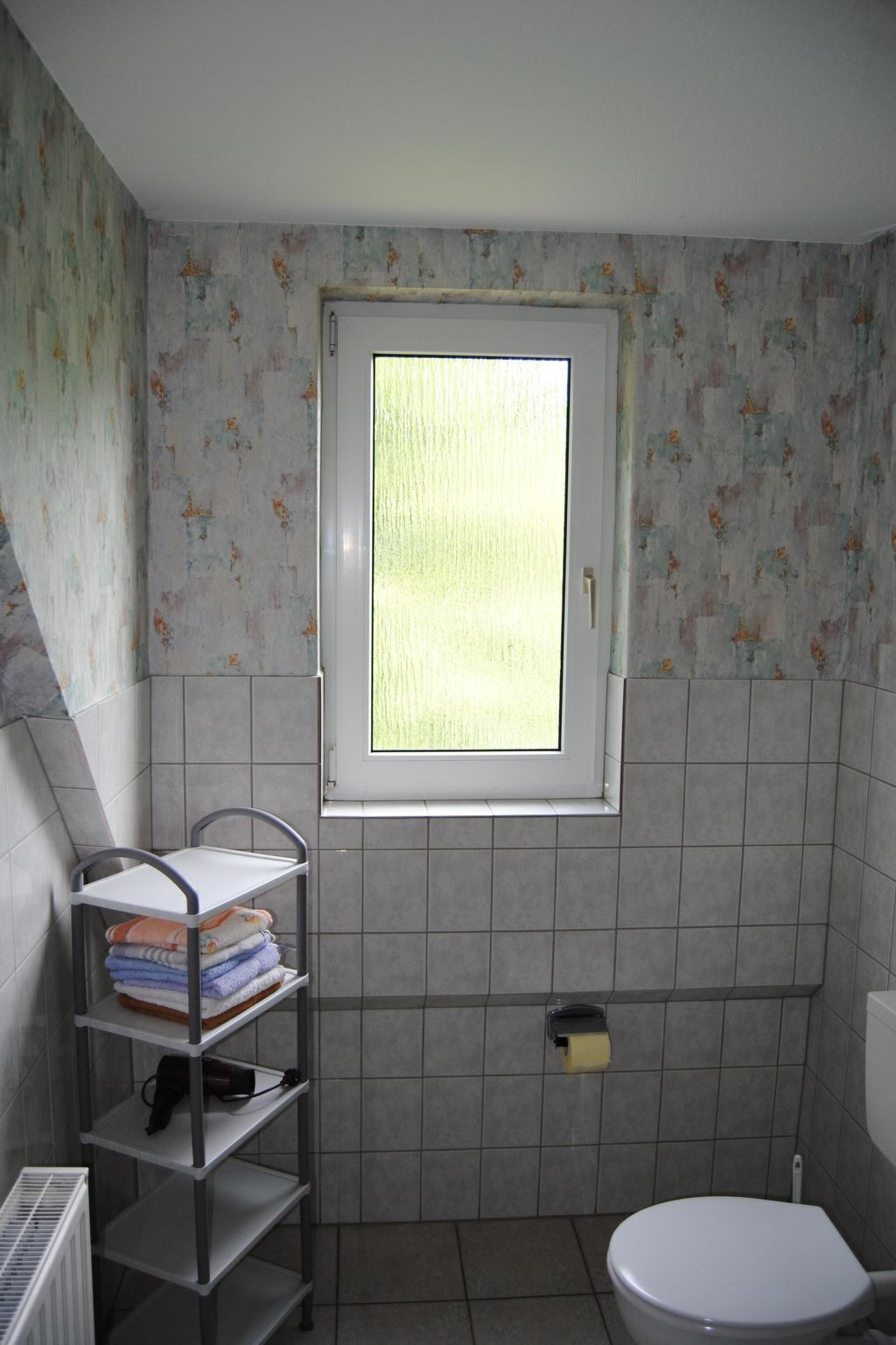 Une petite salle de bain avec une fenêtre, des toilettes et une étagère en métal où sont posées des serviettes.