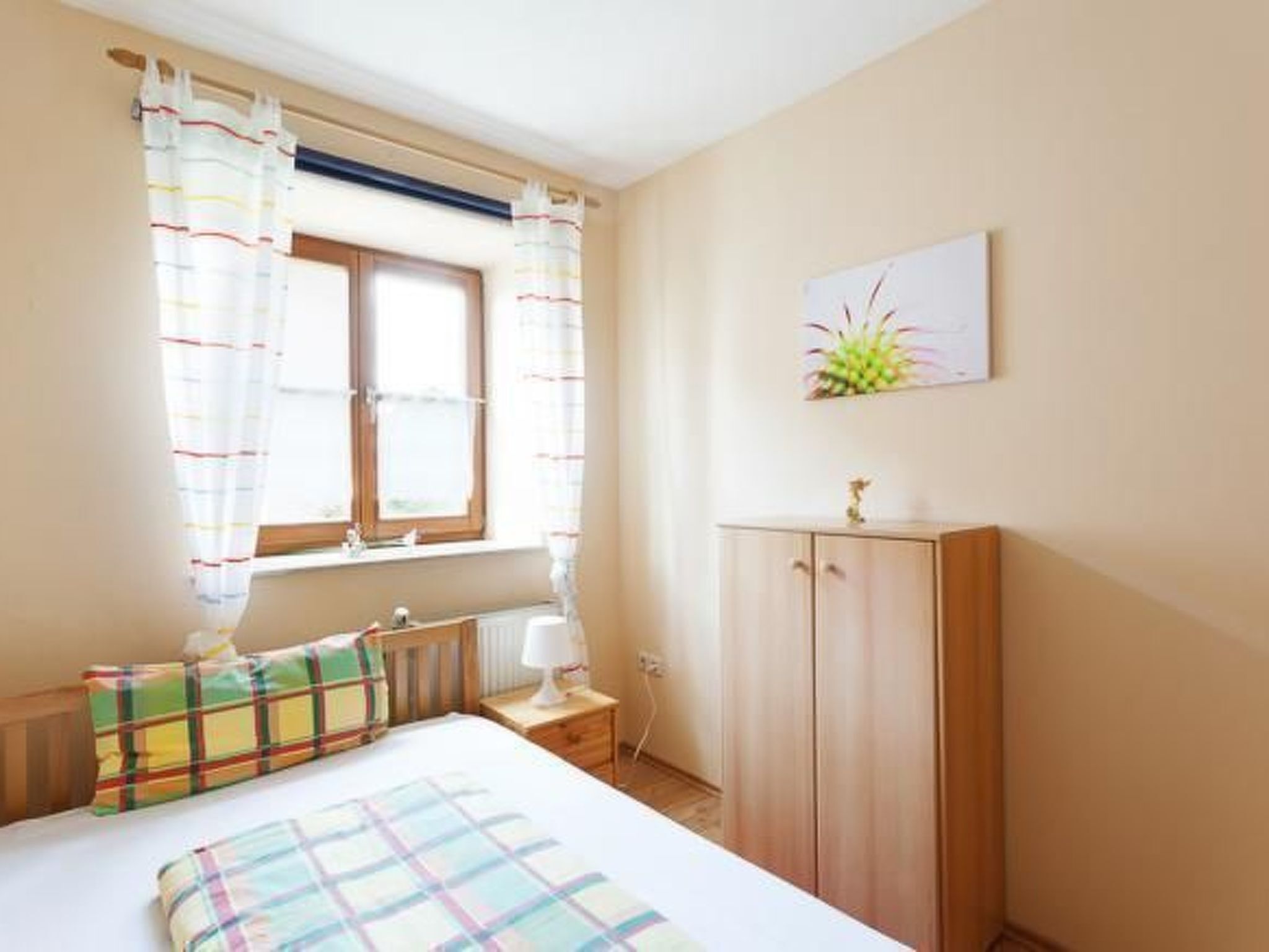 Un dormitorio con cama, mesita de noche, armario de madera, ventana con cortinas y una planta en la pared.
