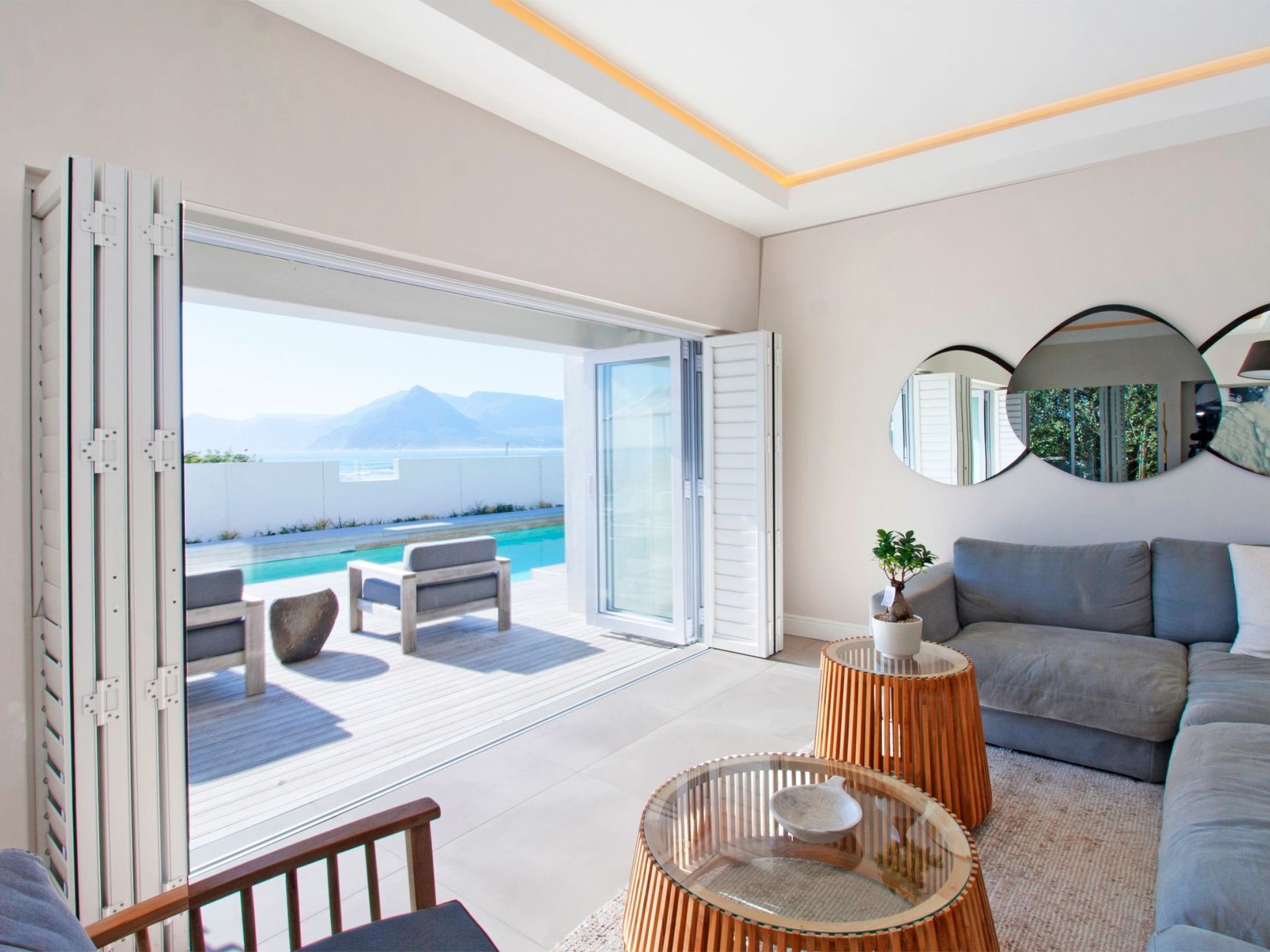 Uma sala de estar moderna com uma grande porta de vidro deslizante que dá para uma área de piscina, um sofá, uma mesa de centro e espelhos decorativos.