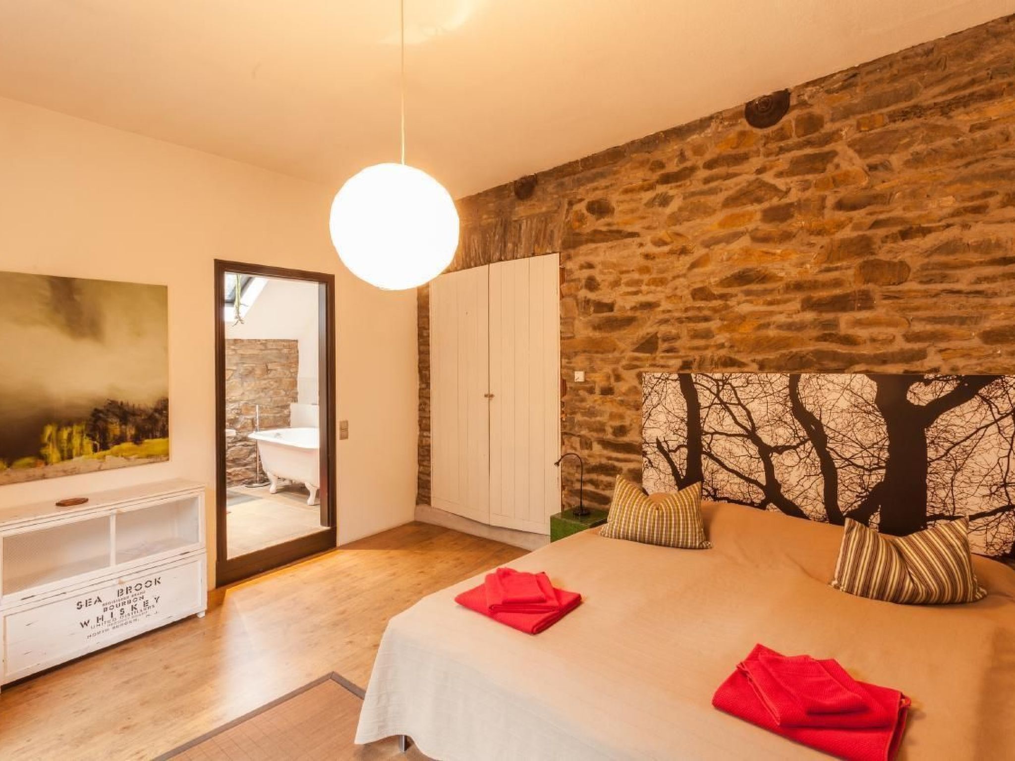 A bedroom with a stone wall, a bed with red towels, a white wardrobe, and a painting on the wall.