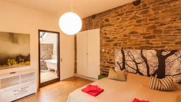 A bedroom with a stone wall, a bed with red towels, a white wardrobe, and a painting on the wall.