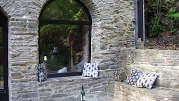 Eine Fassade aus Stein mit Glastür und -fenstern, eine kleine Außenbereich-Sitzgelegenheit mit Kissen und eine Flasche auf einem Glastisch.