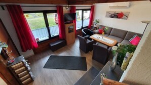 A cozy living room with a sofa, chairs, a coffee table, and a TV. There are windows with red curtains, a wooden bench, and a small table with a vase of flowers. - Vacation home Elster by Interhome (Frielendorf)