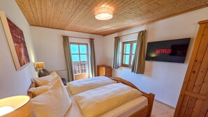 Ein Schlafzimmer mit einem hölzernen Bett, zwei mit Vorhängen versehenen Fenstern, einem an der Wand befestigten Fernseher und einer Holzbalkendecke.