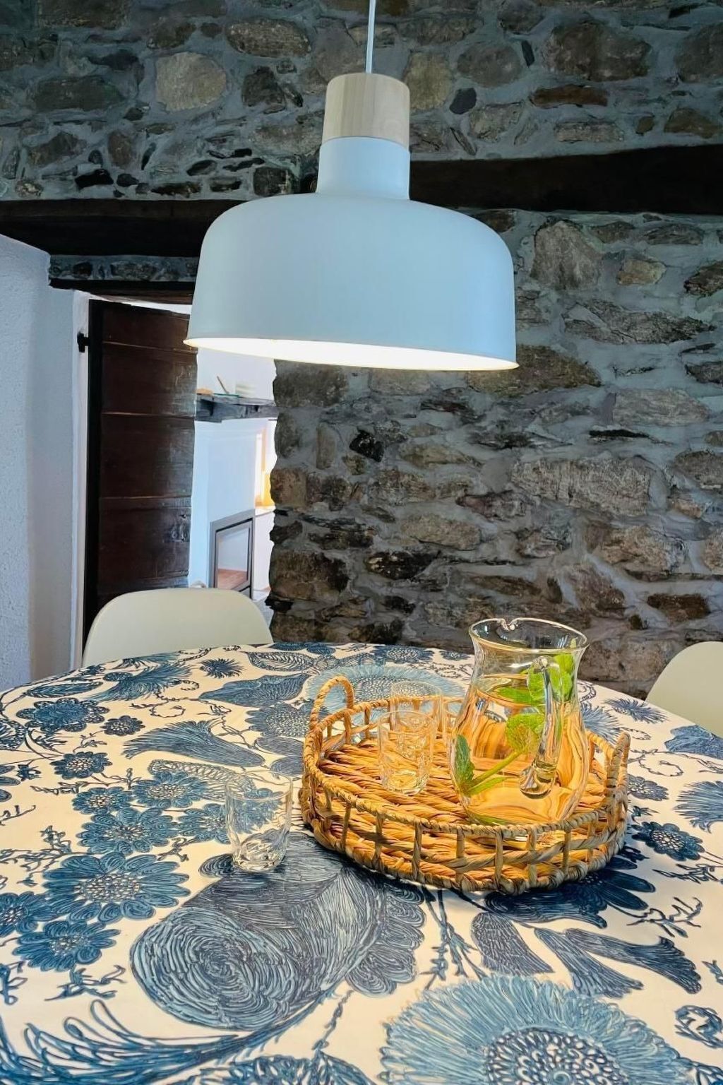 Zona de dinar amb paret de pedra, llum penjant blanca i taula amb mantell blau floral i cistella teixida amb gots i gerra.