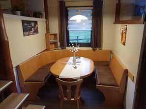 A cozy dining area with a round table and benches, a window with curtains, and a shelf with items. - Vacation home Sonja by Interhome (Frielendorf)