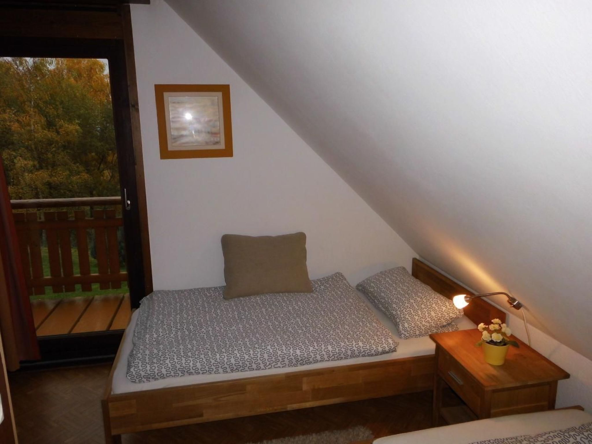 Een knus slaapkamer met een houten bed, een nachtkastje en een kleine tafel met een lamp. Er is een raam met uitzicht op bomen en een houten terras buiten.