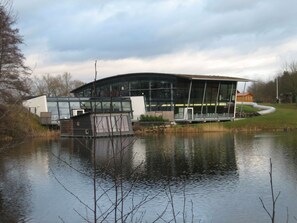 A modern building with large glass windows, a curved roof, and a small dock extending into a calm body of water. - mit Hot Tub by Interhome (Frielendorf)