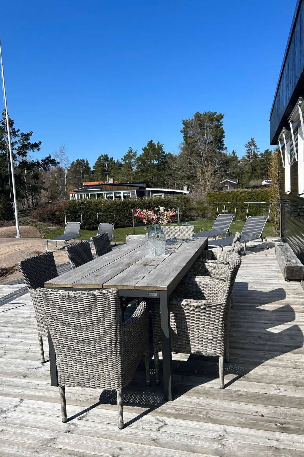 Une terrasse en bois avec une table à manger et des chaises en osier, donnant sur un jardin aménagé et sur un bâtiment situé au loin.