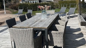 Una terraza de madera con una mesa de comedor y sillas de mimbre, con vistas a un jardín ajardinado y un edificio al fondo.