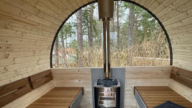 Un cuarto de sauna con bancos de madera, un horno de leña central y vistas a árboles a través de un amplio ventanal de arco.