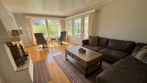 Una sala de estar moderna con piso de madera, un sofá marrón, una mesa de centro y dos sillones. Ventanas amplias ofrecen luz natural y vistas al exterior.