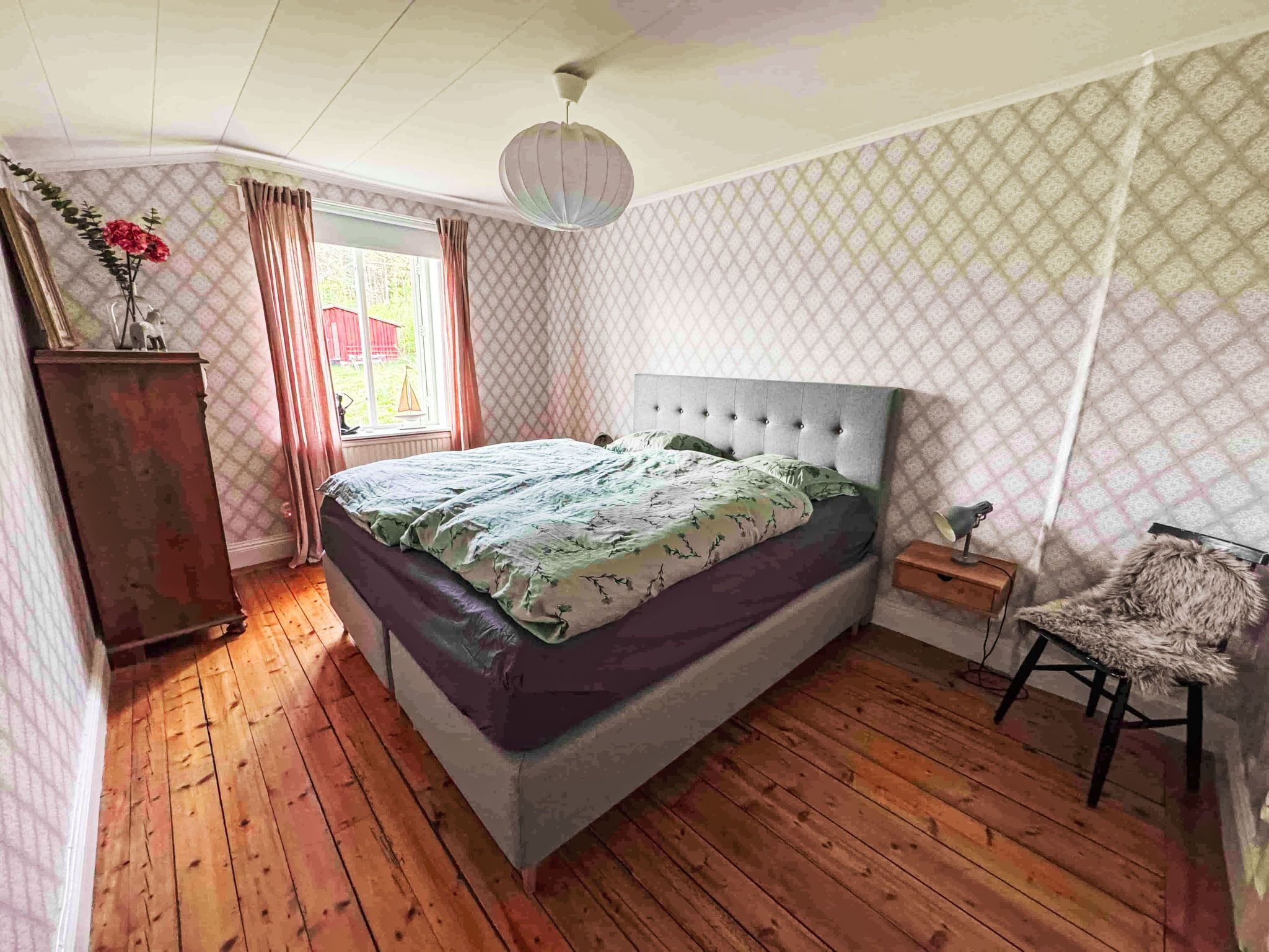 Ein Schlafzimmer mit Holzboden, einem Bett mit gemustertem Überwurf, einem Schminktisch, einem Stuhl und einem Fenster mit Vorhängen.