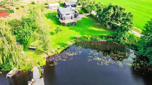 Et stort hus med en terrasse, en sø med siv og en velplejet græsplæne.