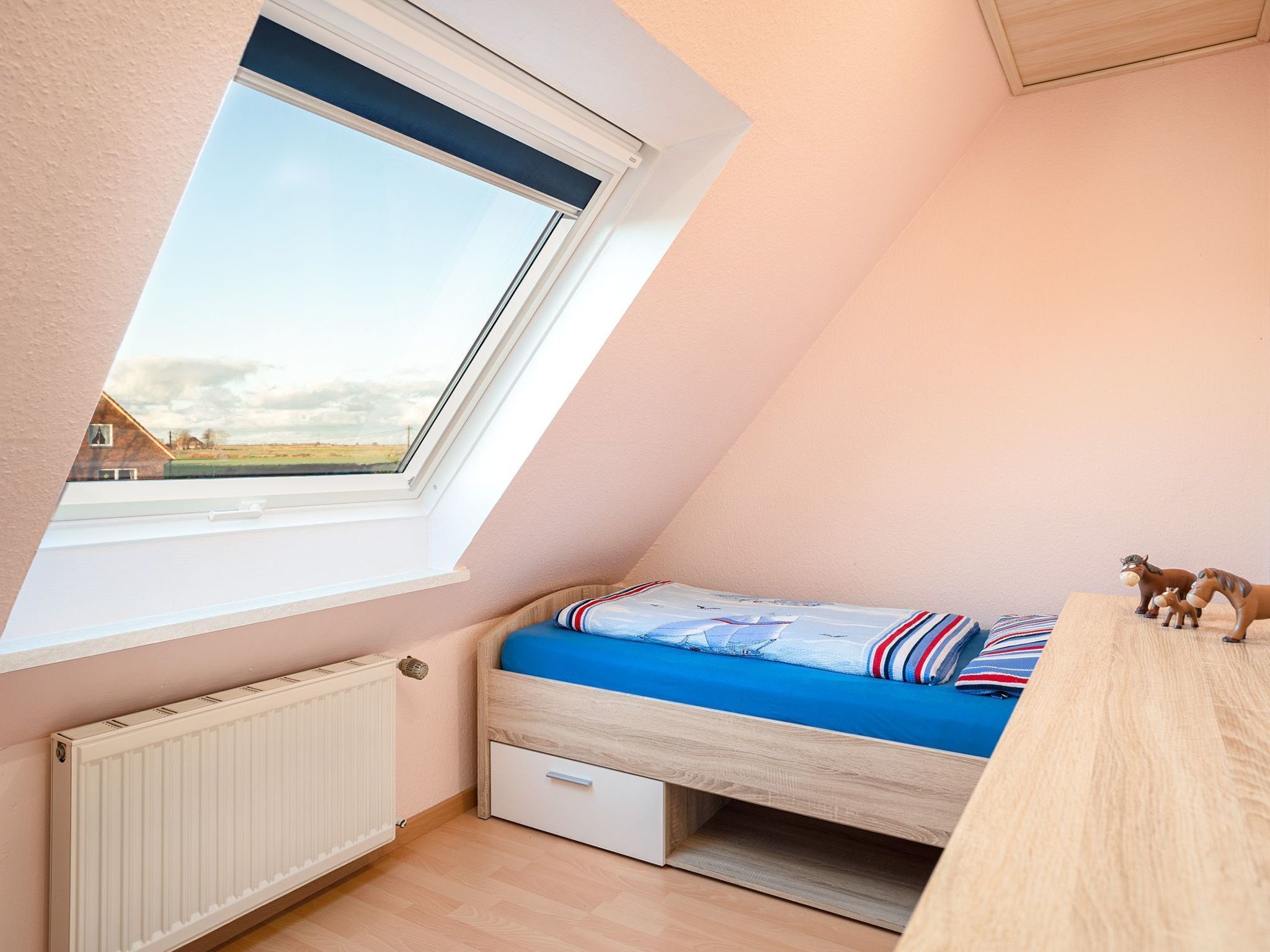 Une petite pièce aux murs inclinés, comprenant un lit simple, un radiateur et une fenêtre donnant sur un paysage rural.
