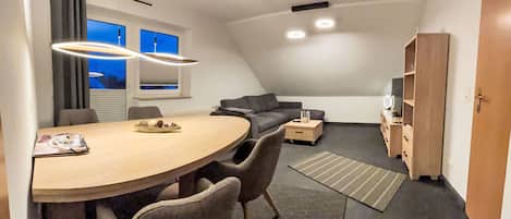 A modern loft-style room with a dining table, chairs, a sofa, a TV, and a bookshelf.
