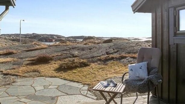 En træhytte med en sten terrasse, en stol og et bord.