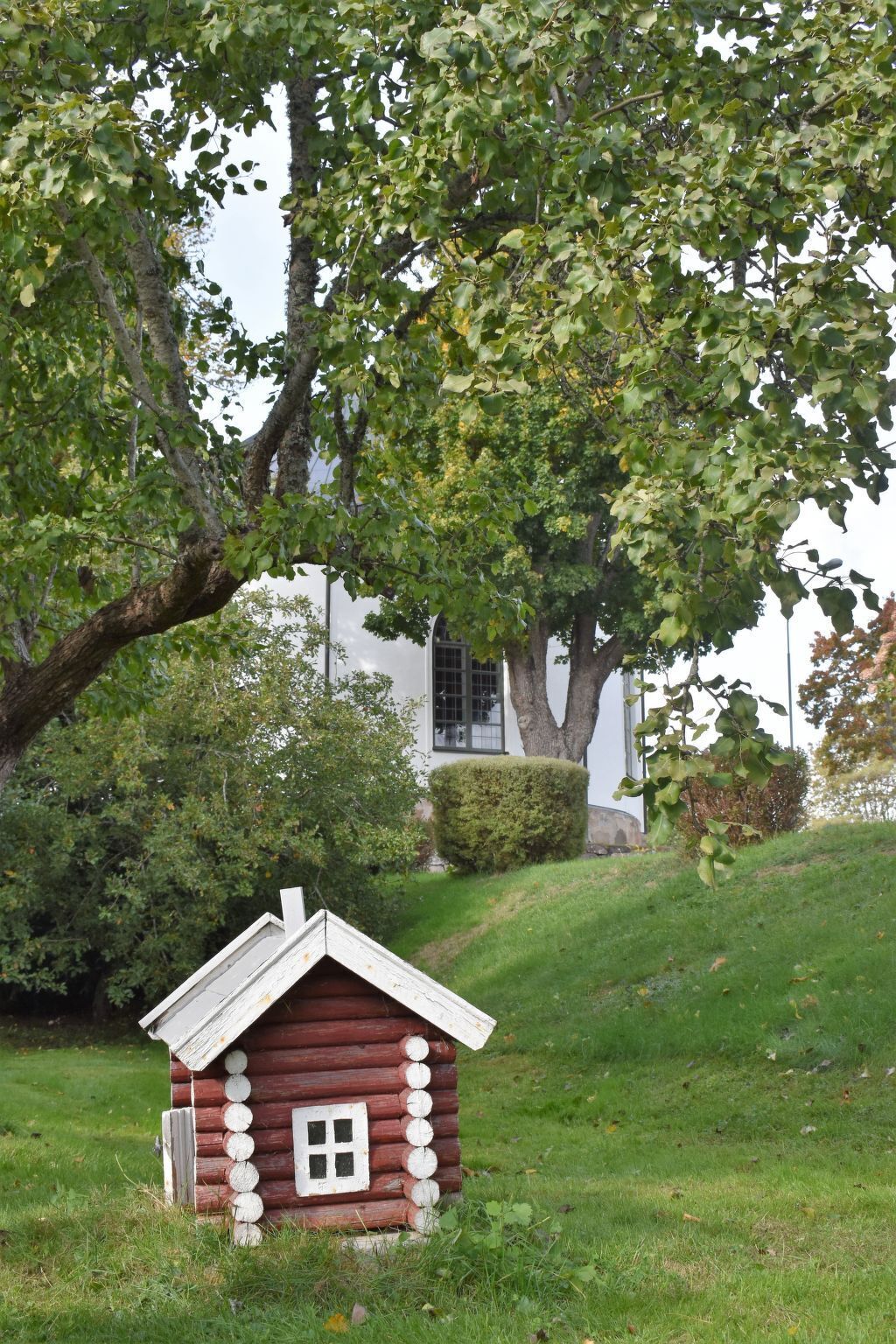Une petite cabane en rondins avec une fenêtre et une porte blanches, entourée d’une végétation luxuriante et d’arbres.