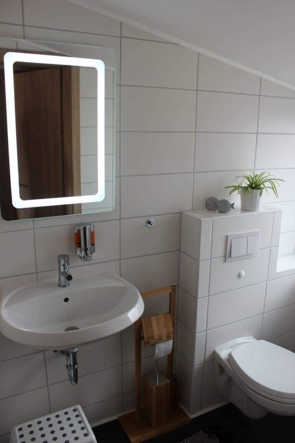 A bathroom with a white sink, mirror, and toilet. There is a small wooden stool and a potted plant on the vanity.