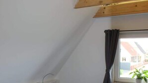 Quarto com teto inclinado, cama com roupa de cama listrada em azul e branco, radiador e janela com cortinas.