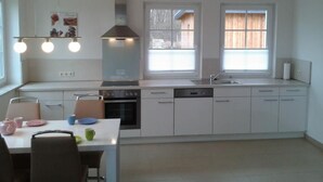 Eine moderne Küche mit weißen Schränken, einem Herd, Backofen und Geschirrspüler. Zwei Fenster mit Jalousien, eine Deckenleuchte sowie ein kleiner Essbereich mit Tisch und Stühlen.