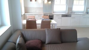 Ein modernes Wohnzimmer mit einem grauen Ecksofa, einem Esstisch mit Stühlen und einer Küche im Hintergrund.