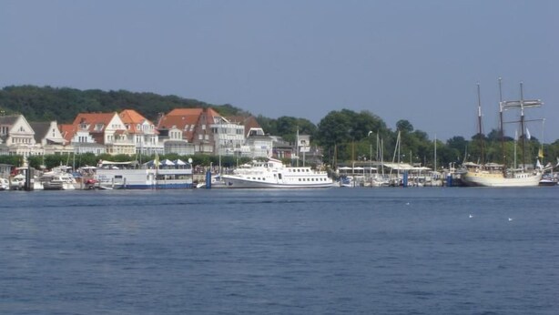 Ein Hafen mit einem großen weißen Boot, das an einem Yachthafen liegt, umgeben von Gebäuden mit roten Dächern und einem bewaldeten Hügel im Hintergrund.
