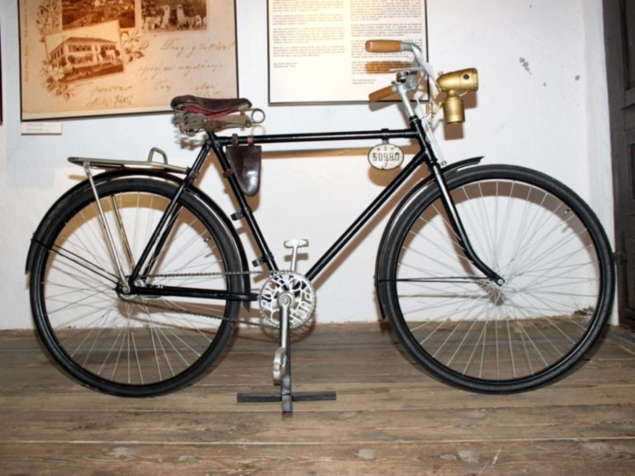 Una bicicleta antigua con sillín de cuero y una campanilla de bronce, exhibida sobre un suelo de madera frente a una pared con un documento enmarcado.