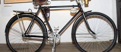 Una bicicleta antigua con sillín de cuero y una campanilla de bronce, exhibida sobre un suelo de madera frente a una pared con un documento enmarcado.