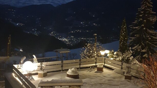 Ein verschneiter Außenbereich mit Bänken und einem kleinen Tisch, umgeben von schneebedeckten Bäumen und Bergen im Hintergrund.