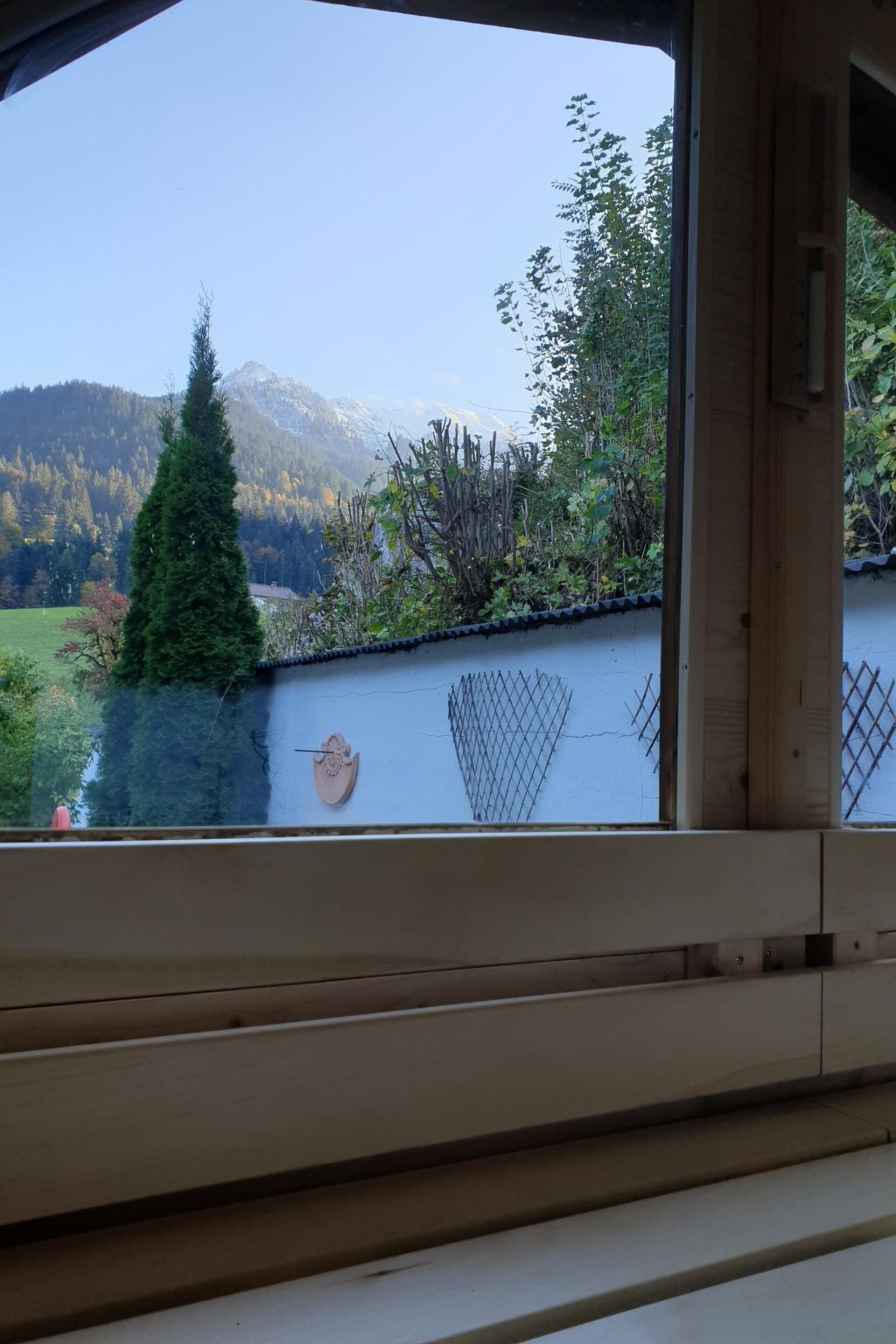 A view from inside a room with a window overlooking a mountain landscape, a wooden fence, and a small tree.