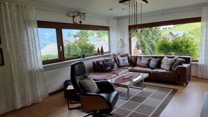 Ein Wohnzimmer mit einer braunen Ledersofa, einem schwarzen Sessel und einem Couchtisch. Große Fenster mit weißen Vorhängen, Blick ins Grüne und eine Deckenleuchte sind ebenfalls vorhanden.