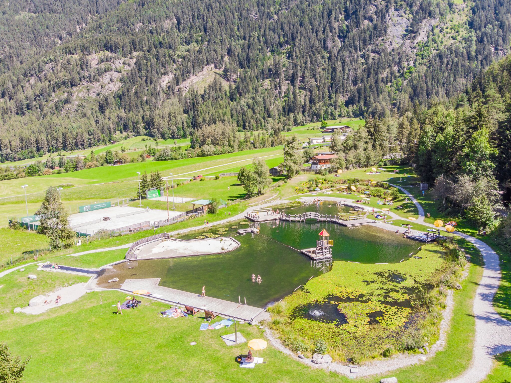 An aerial view of a lakeside park with a wooden dock, a small pavilion, and a grassy area with people sunbathing.