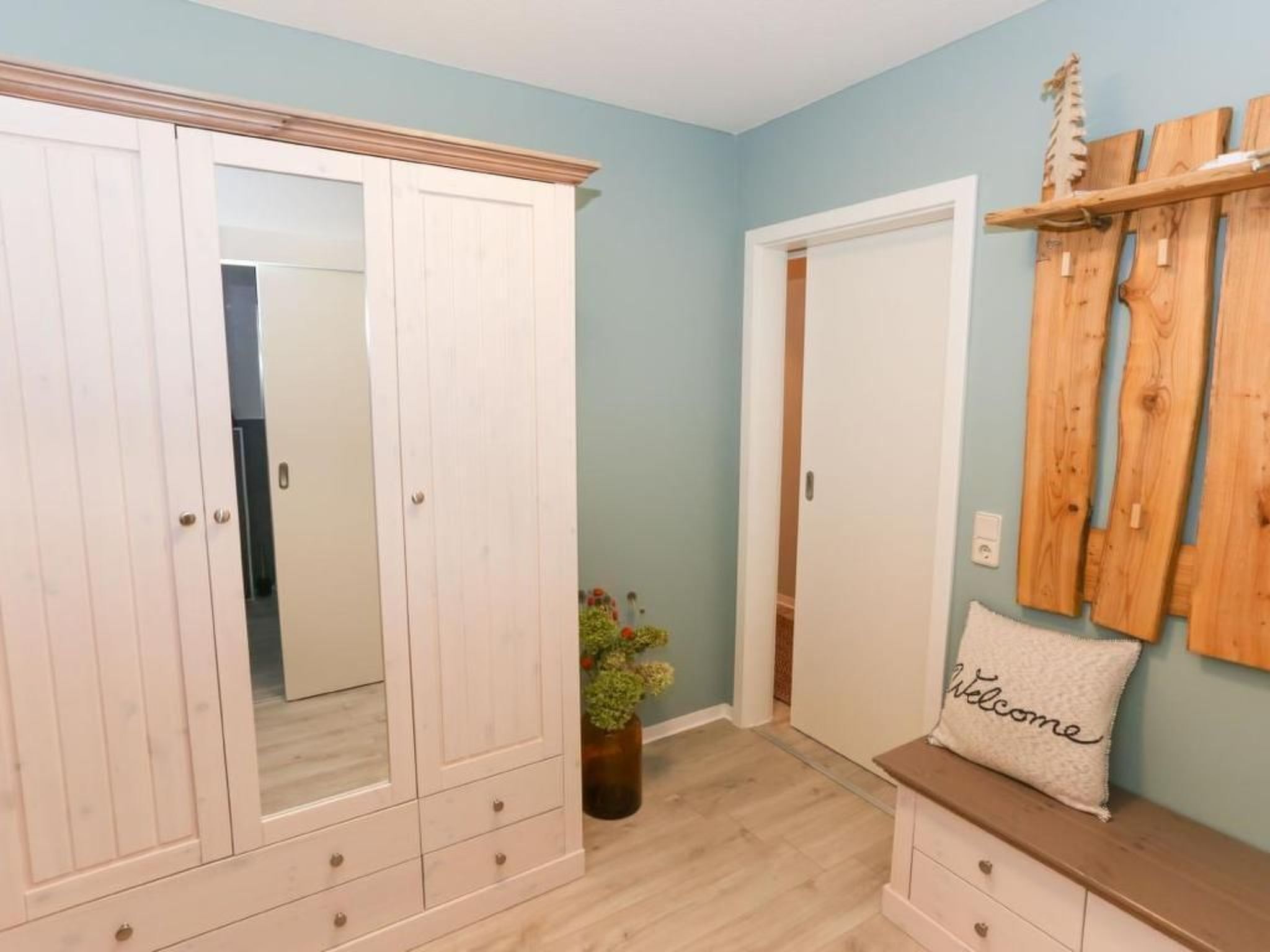 Ein Zimmer mit einem großen weißen Kleiderschrank, einer Holzbank mit einem 'Welcome'-Kissen und einem Holzregal mit einer kleinen Pflanze.