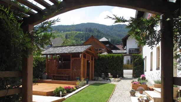 A garden with a wooden pergola, a wooden deck, and a gravel path leading to a house with a sloped roof. - Chalet Gove für Unterkunft mit Halbpension unter der Leitung des Eigentümers by Interhome (Tamsweg im Lungau)