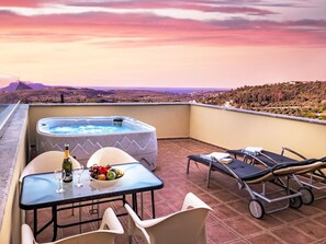 Un balcon avec un jacuzzi, une table avec des verres à vin et des fruits, et des chaises longues.