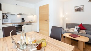 Cocina moderna con armarios blancos, microondas y cafetera. Hay una mesa de comedor de madera con copas de vino y frascos. Un sofá gris con un cojín rojo se encuentra en la sala de estar.