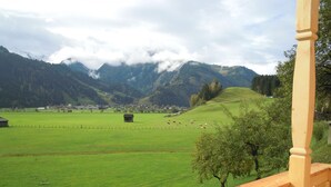 Vista desde un balcón de madera con un campo verde, vacas, un edificio pequeño y montañas al fondo.